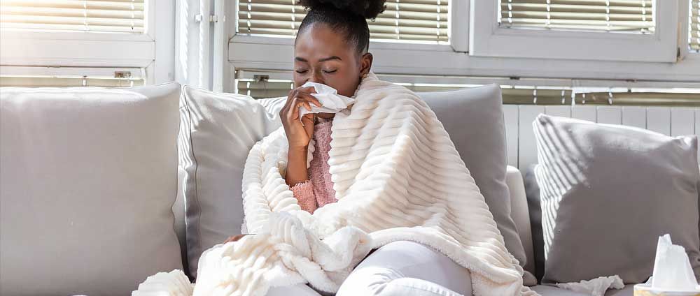 A woman sitting on a couch wrapped in a blanket, sneezing into a tissue, appearing to have a cold or flu